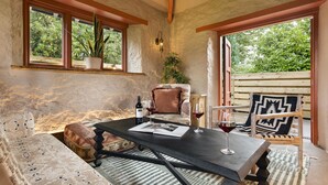 A cozy room with a stone wall, a wooden ceiling, a black table with a book and wine glasses, a cushioned bench, and a patterned chair. - Richmond Place - Apartment in Private Mansion (Kendal)
