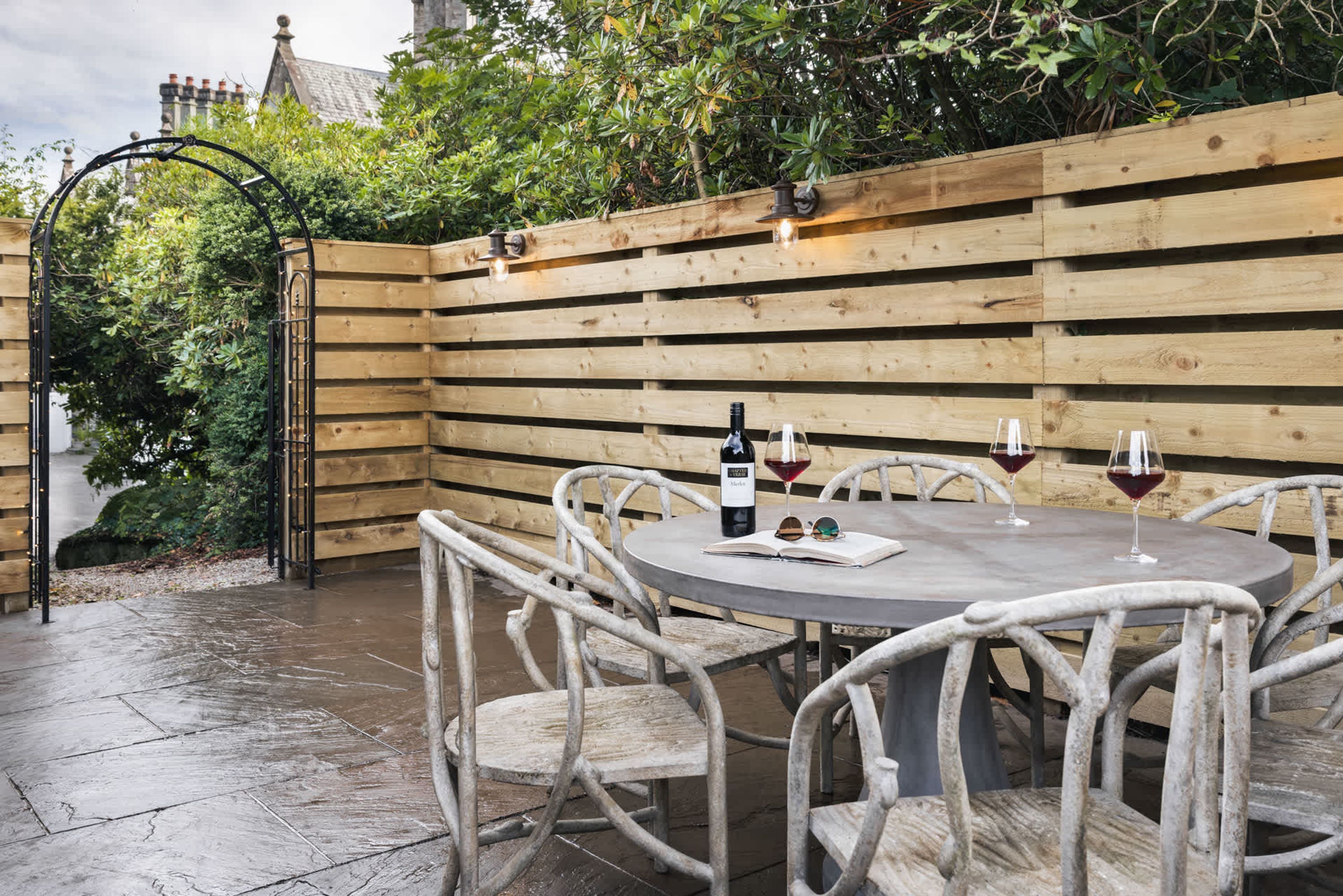A round table with chairs, a bottle of wine, and glasses, set against a wooden fence with a metal gate.