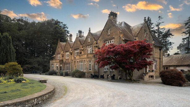 A traditional stone building with multiple gabled roofs, a gravel driveway, and a well-maintained garden with a prominent red flowering tree. - Richmond Place - Apartment in Private Mansion (Kendal)