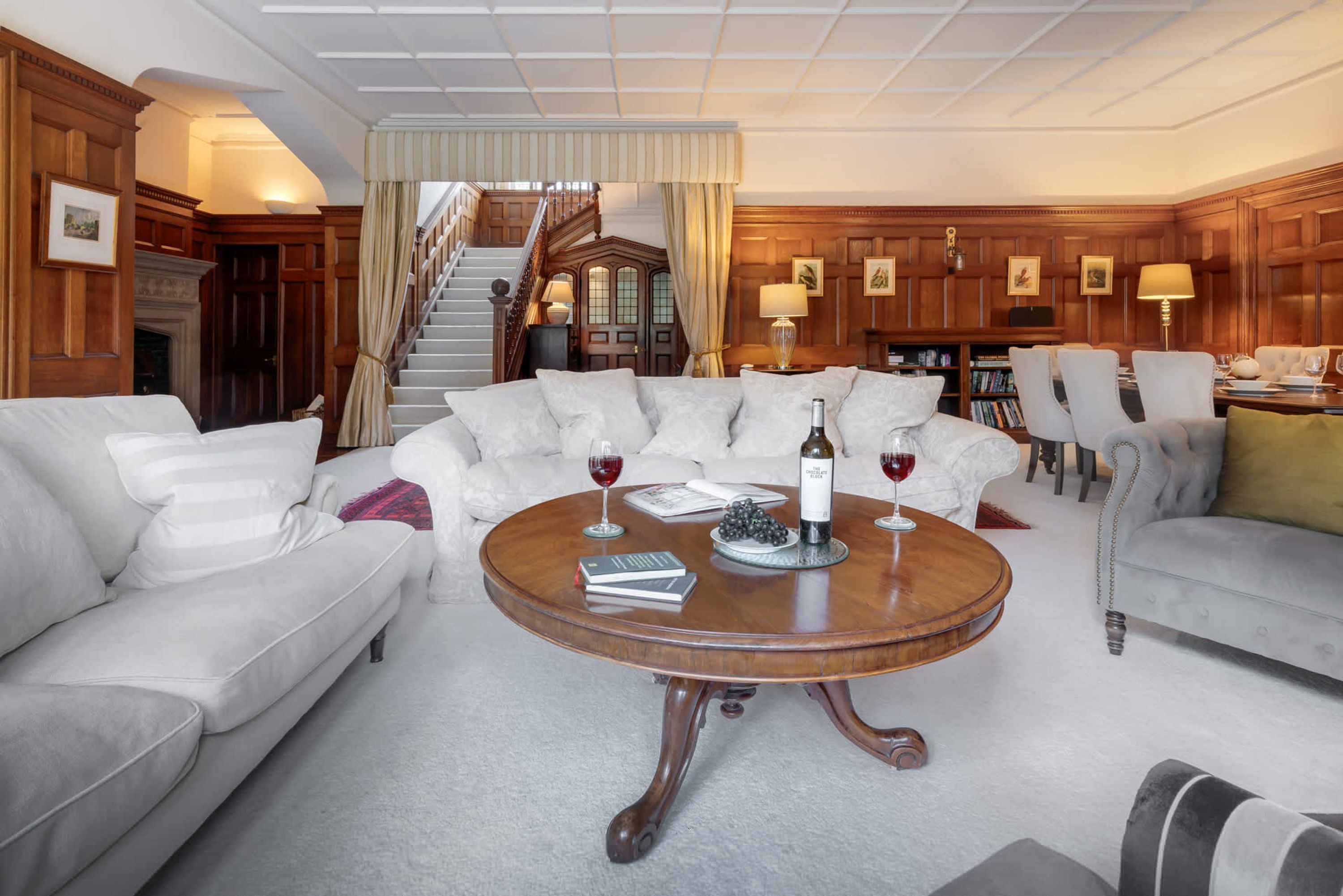 Een ruime woonkamer met een ronde houten salontafel, witte banken en een trap naar een hoger gelegen verdieping.