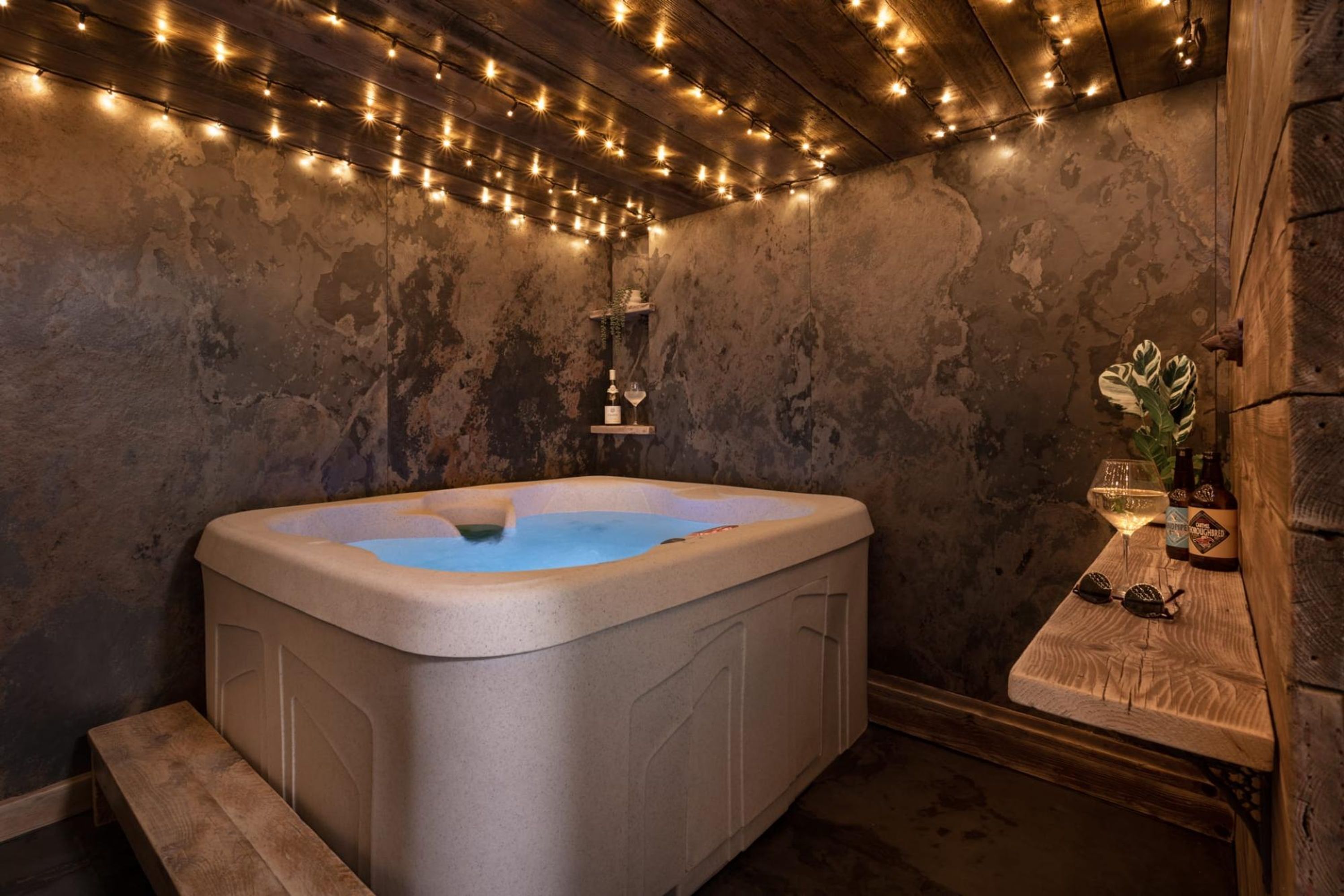 Un jacuzzi con un banco de madera, un estante con una copa y una botella de vino, y luces colgantes en el techo.