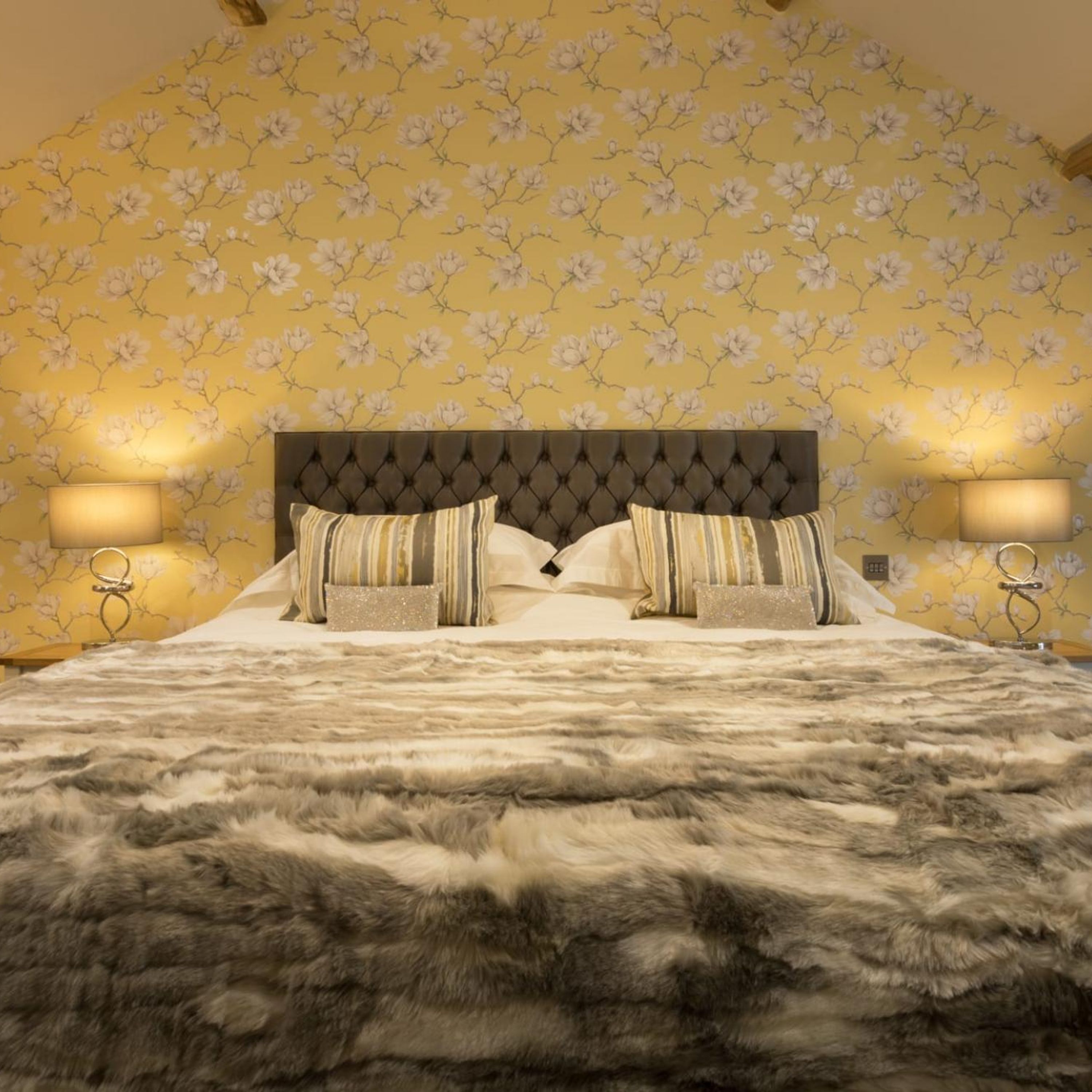 A bedroom with a tufted headboard, two beds with striped bedding, a fluffy rug, and floral wallpaper.