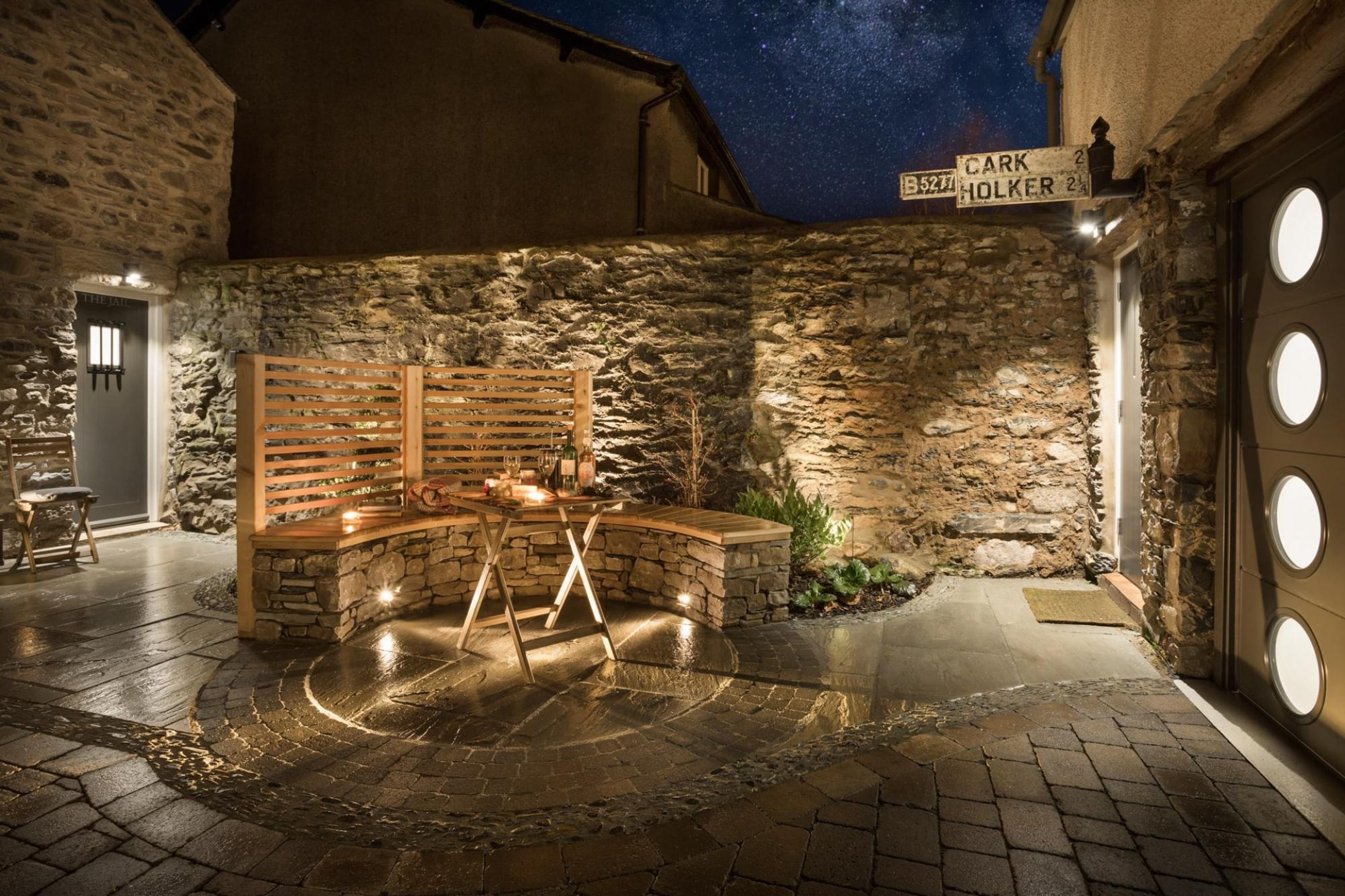 A stone-walled outdoor area with a wooden bench, a small table with candles, and a sign indicating 'CARK HOLKER'.