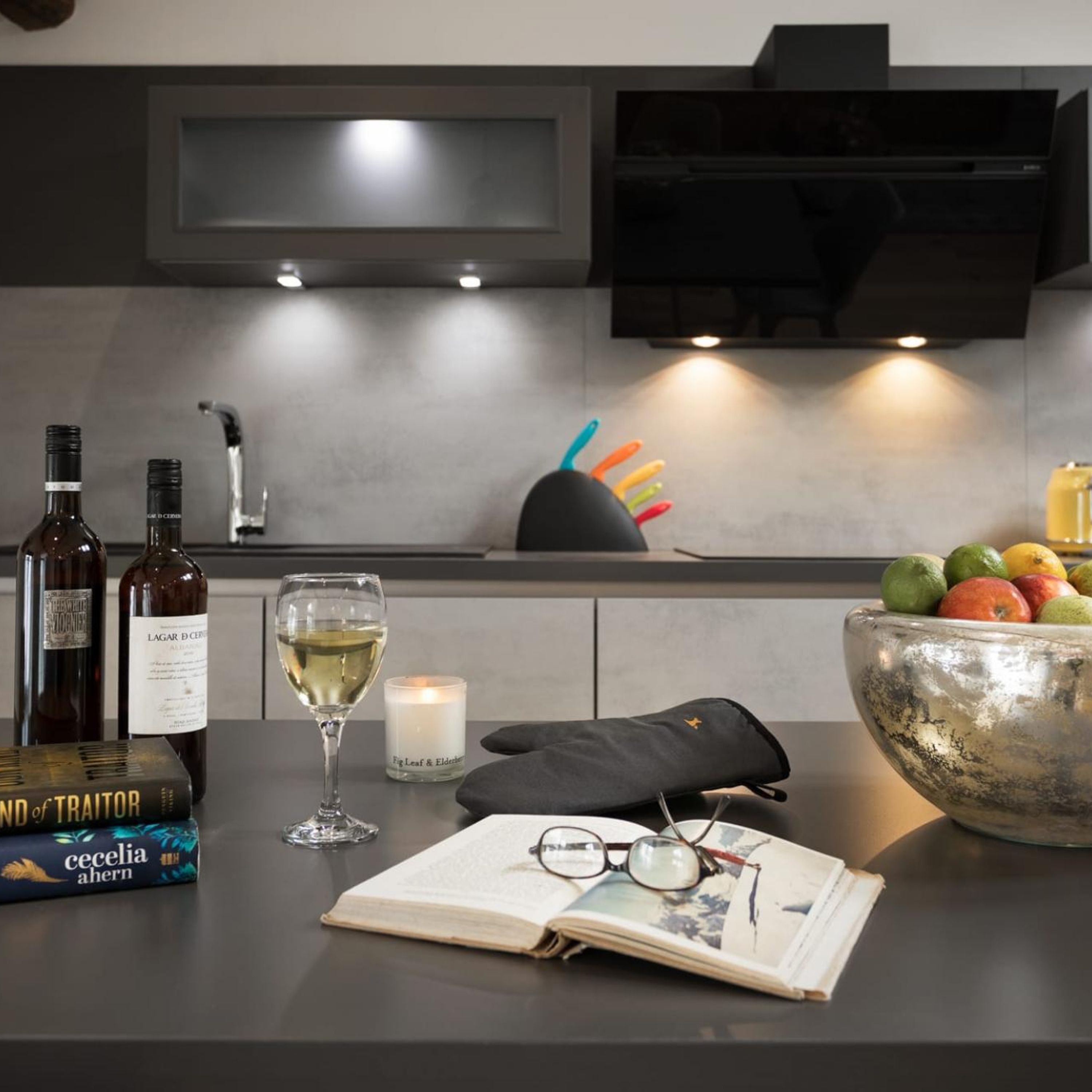 A modern kitchen with a wine bottle, a glass of white wine, a book, glasses, a candle, and a fruit bowl.