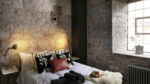 A bedroom with a stone wall, a bed with black and white patterned pillows, a red pillow, and a bedside table with a lamp. - Two-bedroom Apartment at Royal William Yard (Plymouth)