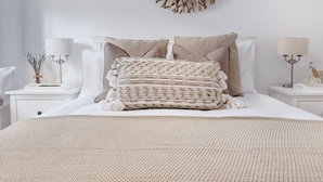 Una cama bien hecha con una colcha color beige y almohadas, un adorno en la pared y lámparas de noche.