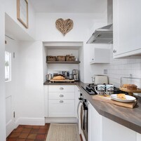 Una cocina compacta con armarios blancos, una decoración en forma de corazón en la pared, una encimera con hogaza de pan, una cafetera y un plato con un sándwich.