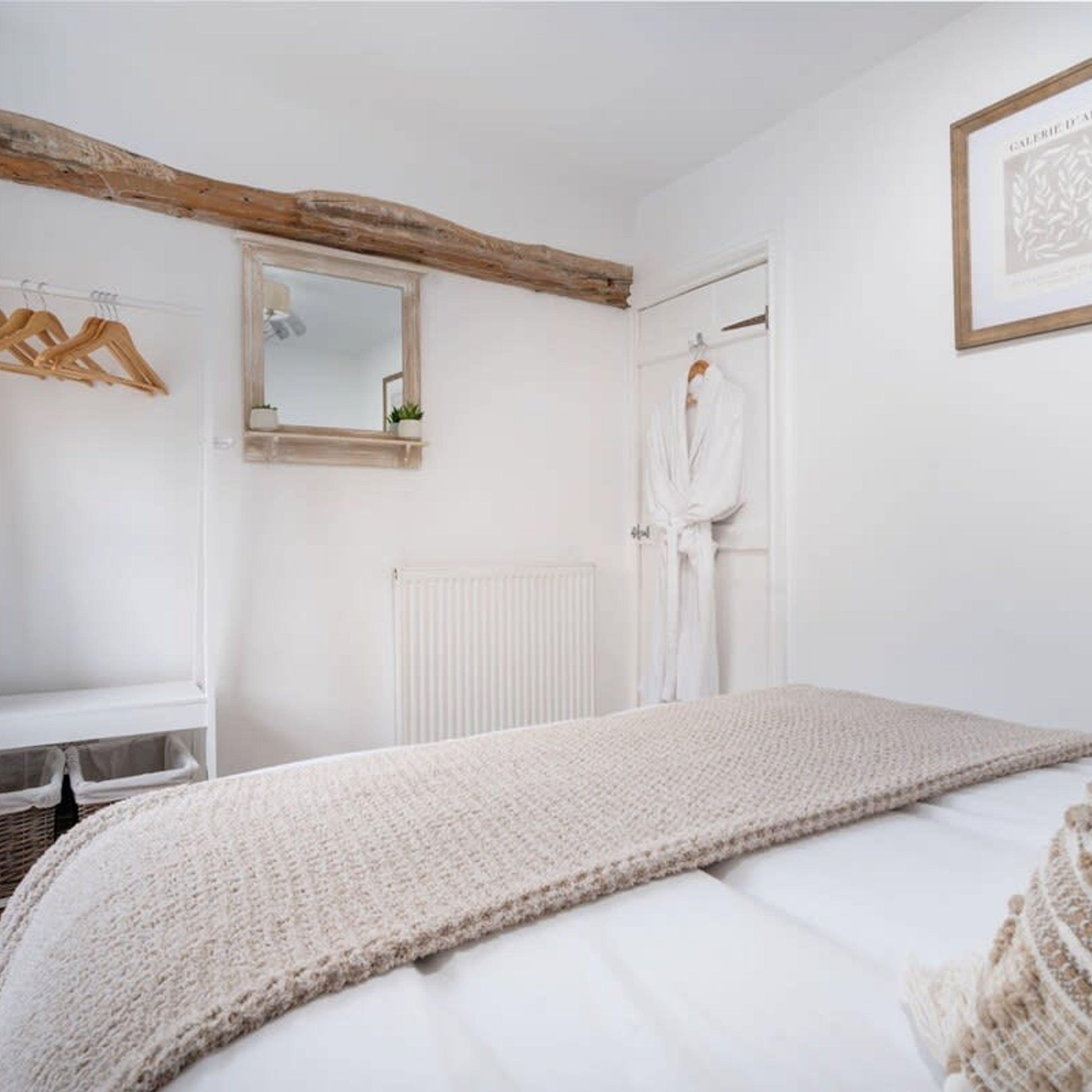 A bedroom with a bed, a wooden beam ceiling, a white radiator, a mirror, and a framed picture on the wall.