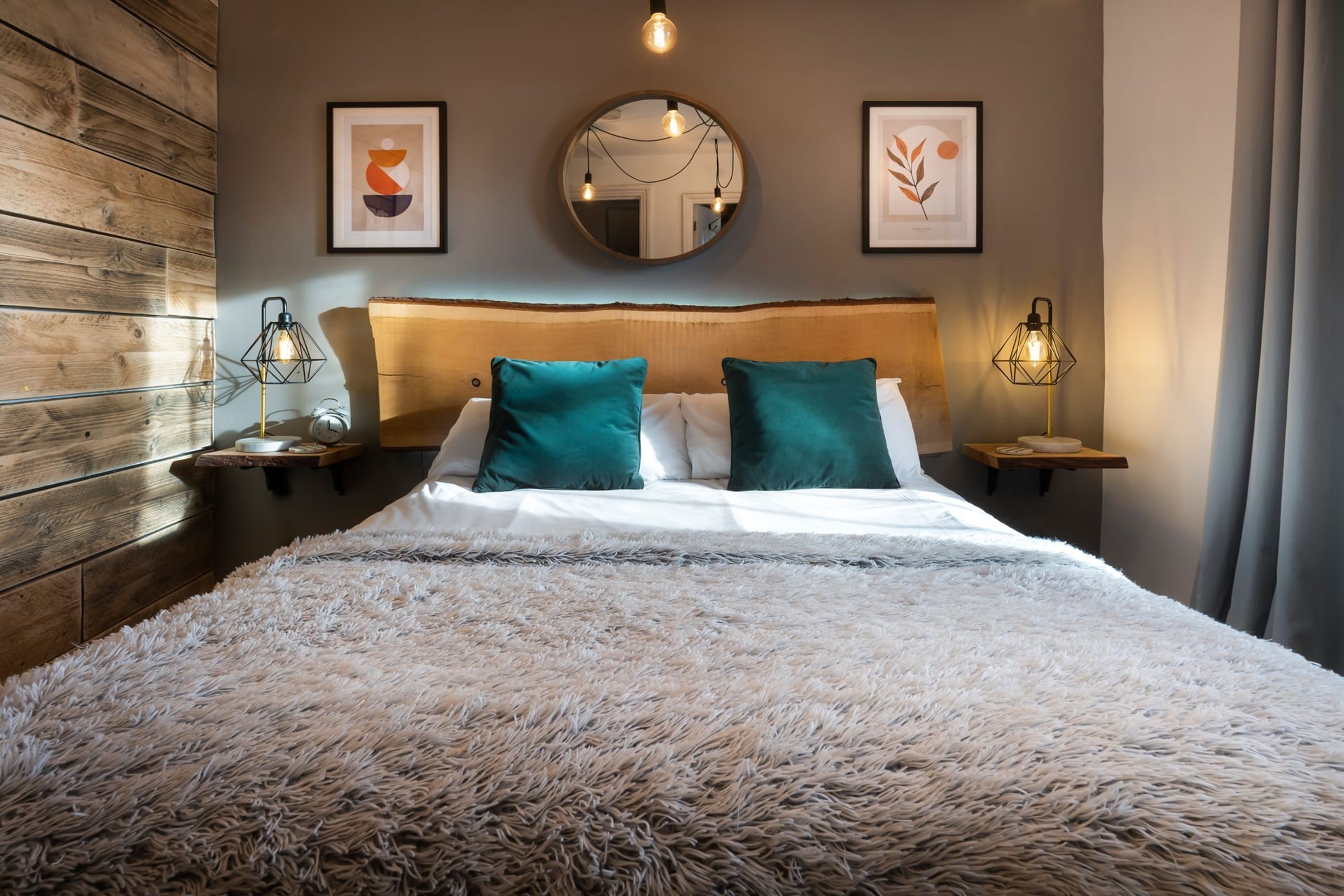 Une chambre avec une tête de lit en bois, une couette blanche moelleuse, des coussins turquoise, un miroir rond et deux lampes fixées au mur.