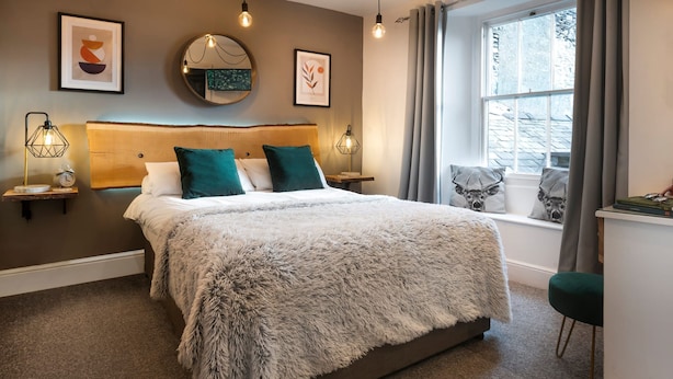 Une chambre à coucher moderne avec un grand lit, une tête de lit, des lampes de chevet, une fenêtre avec des rideaux et des œuvres d’art accrochées aux murs.