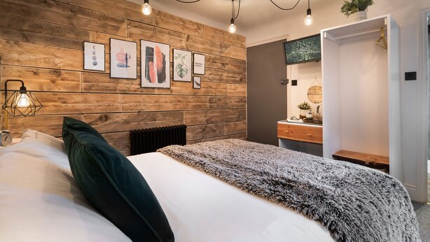 A bedroom with a wooden headboard, a bed with white and dark green bedding, a fluffy grey throw blanket, and a bedside table with a lamp. - Windermere Central Hotel - Room 1 (Windermere)
