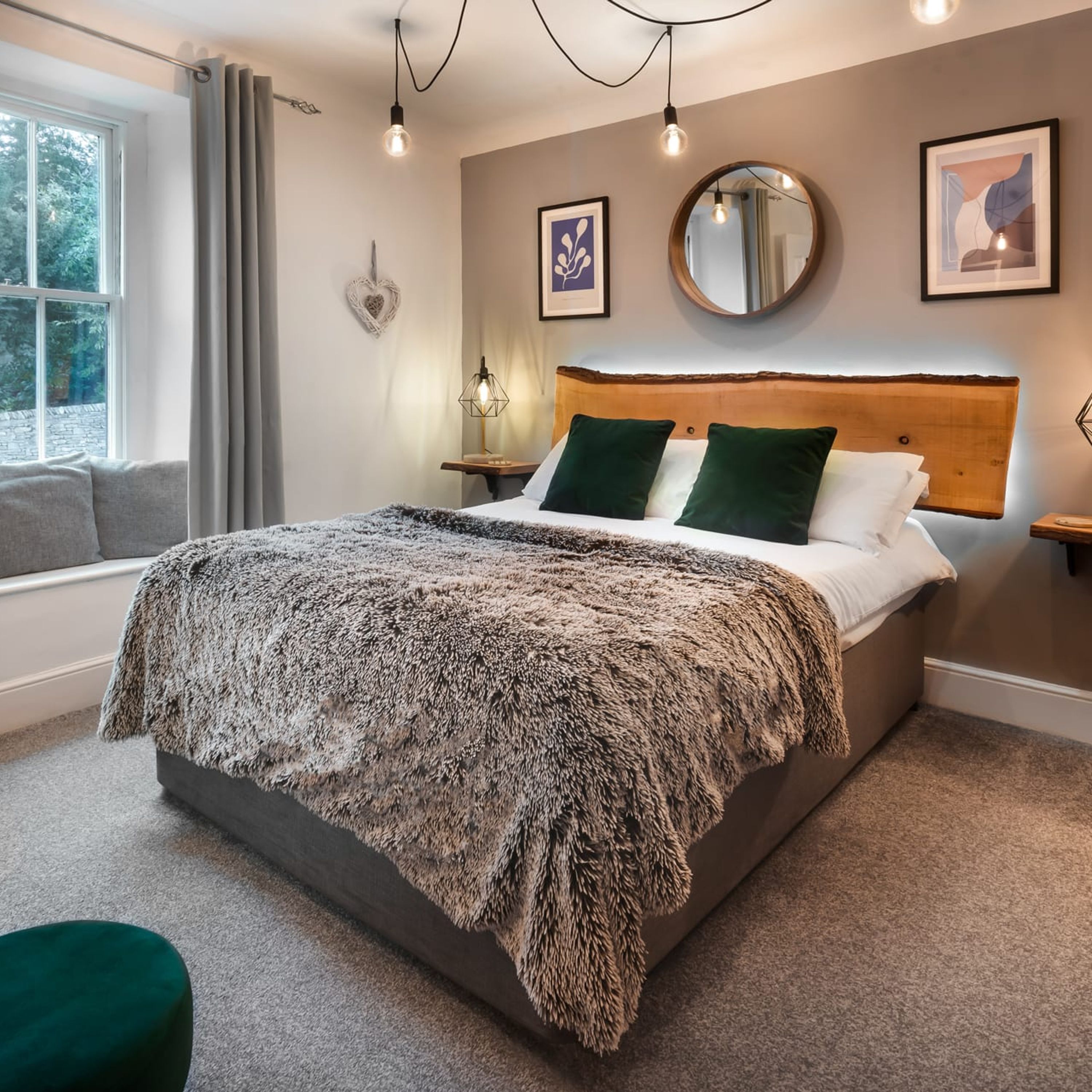 A bedroom with a large bed, a window with curtains, a bedside lamp, and a wall-mounted mirror.