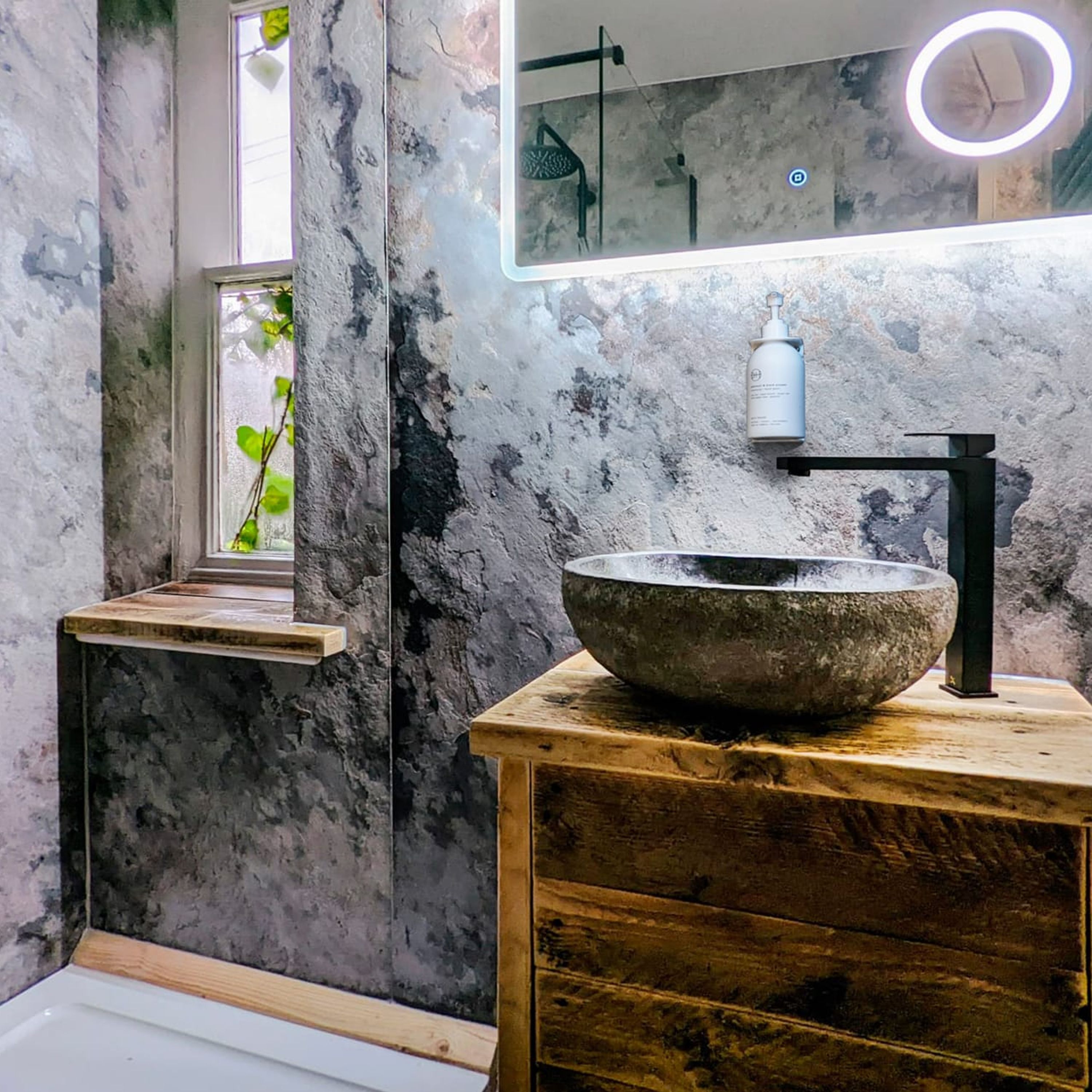 A modern bathroom with a large stone sink, a wooden vanity, a mirror with a circular light, and a window with greenery.