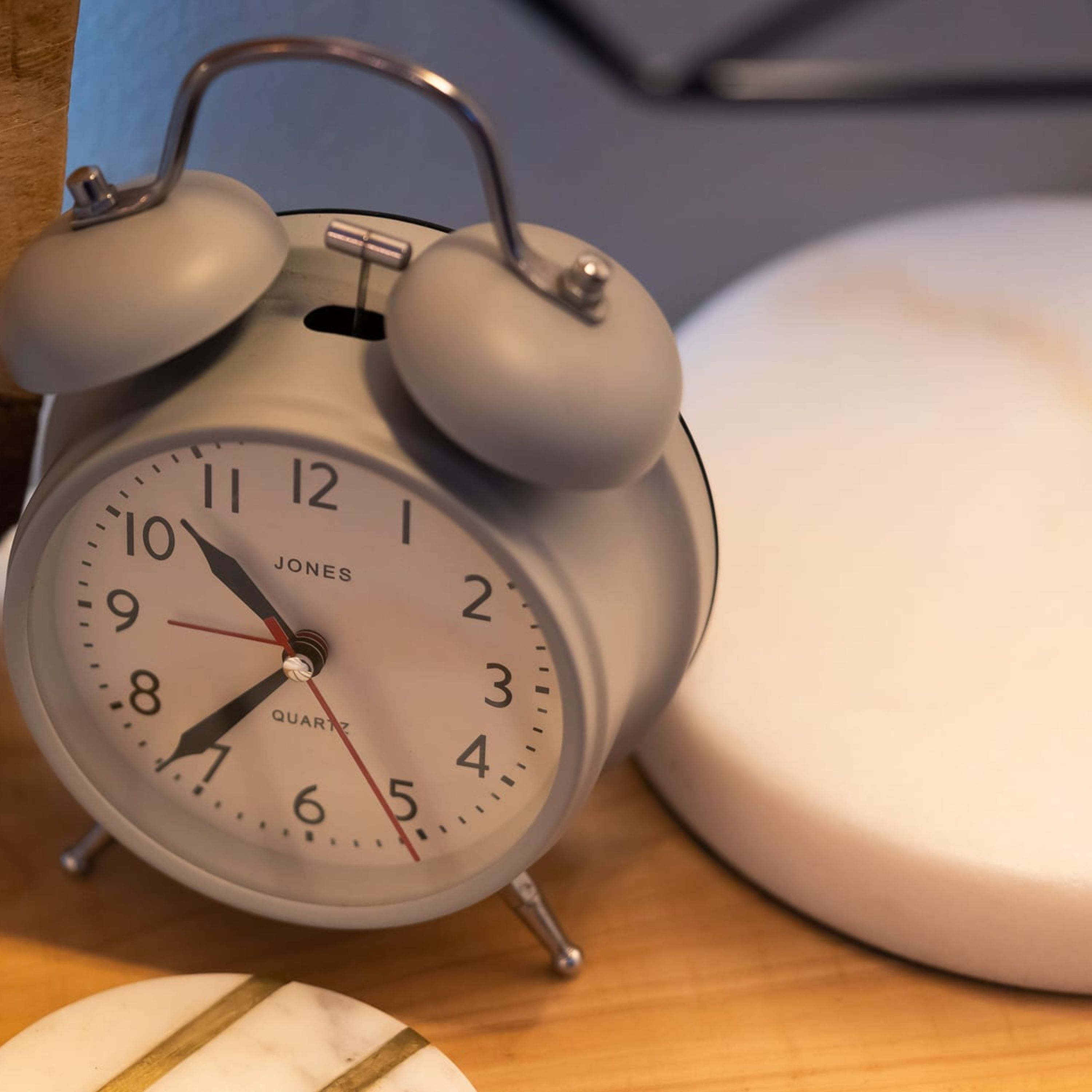 Un reloj despertador Jones antiguo de cuarzo, con esfera blanca y números negros, sobre una superficie de madera junto a un objeto blanco.