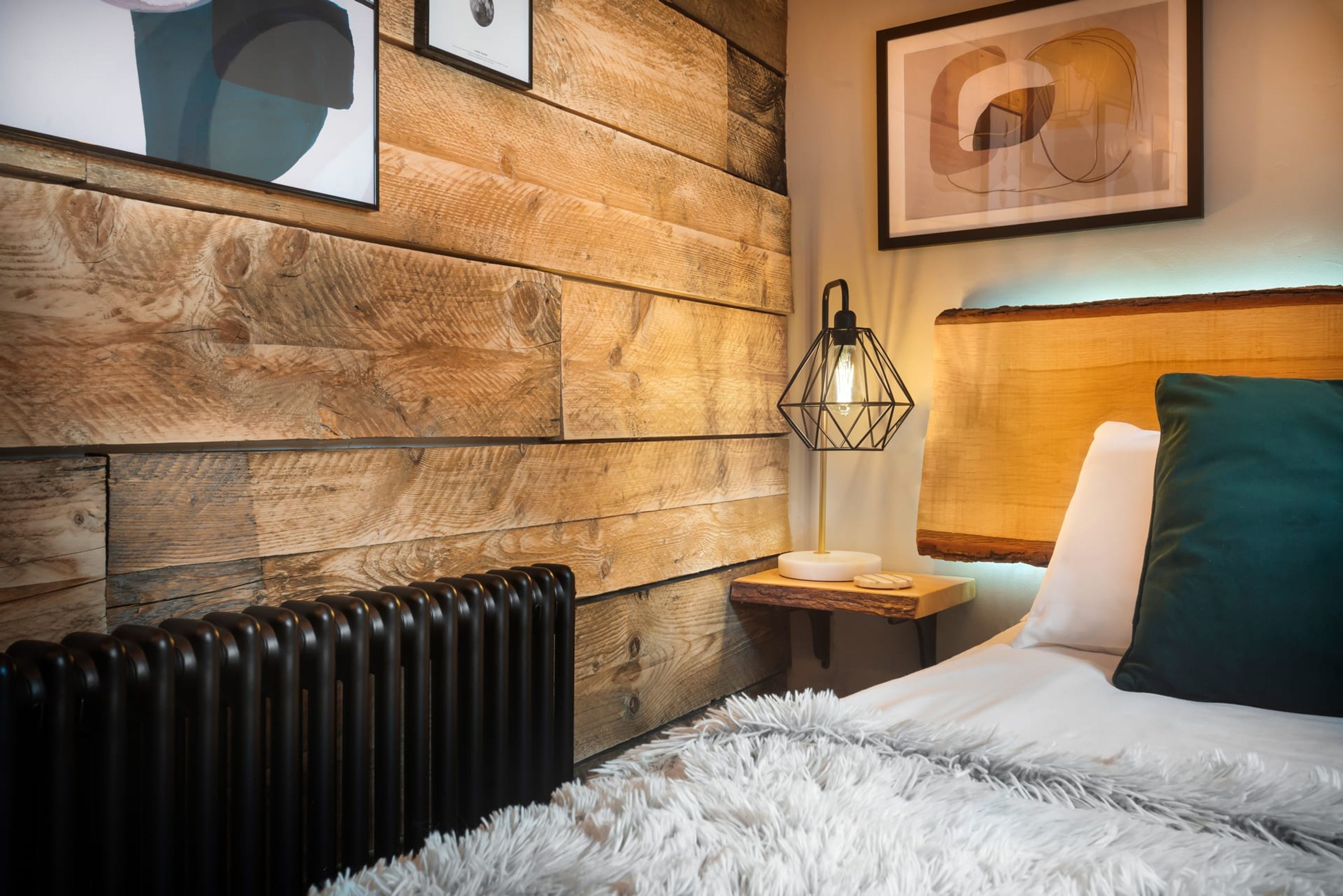 A bedroom with a wooden wall panel, a radiator, a fluffy white rug, a bedside table with a lamp, and a bed with a green pillow.