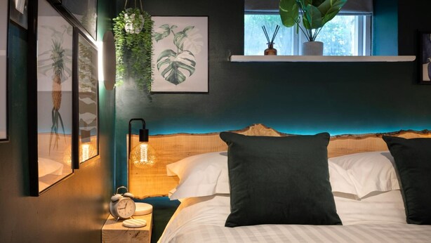 A bedroom with a bed, pillows, a nightstand, a clock, wall art, and a window with plants. - Eden Escape - Comfy Apartment Quiet Neighbourhood (Kendal)