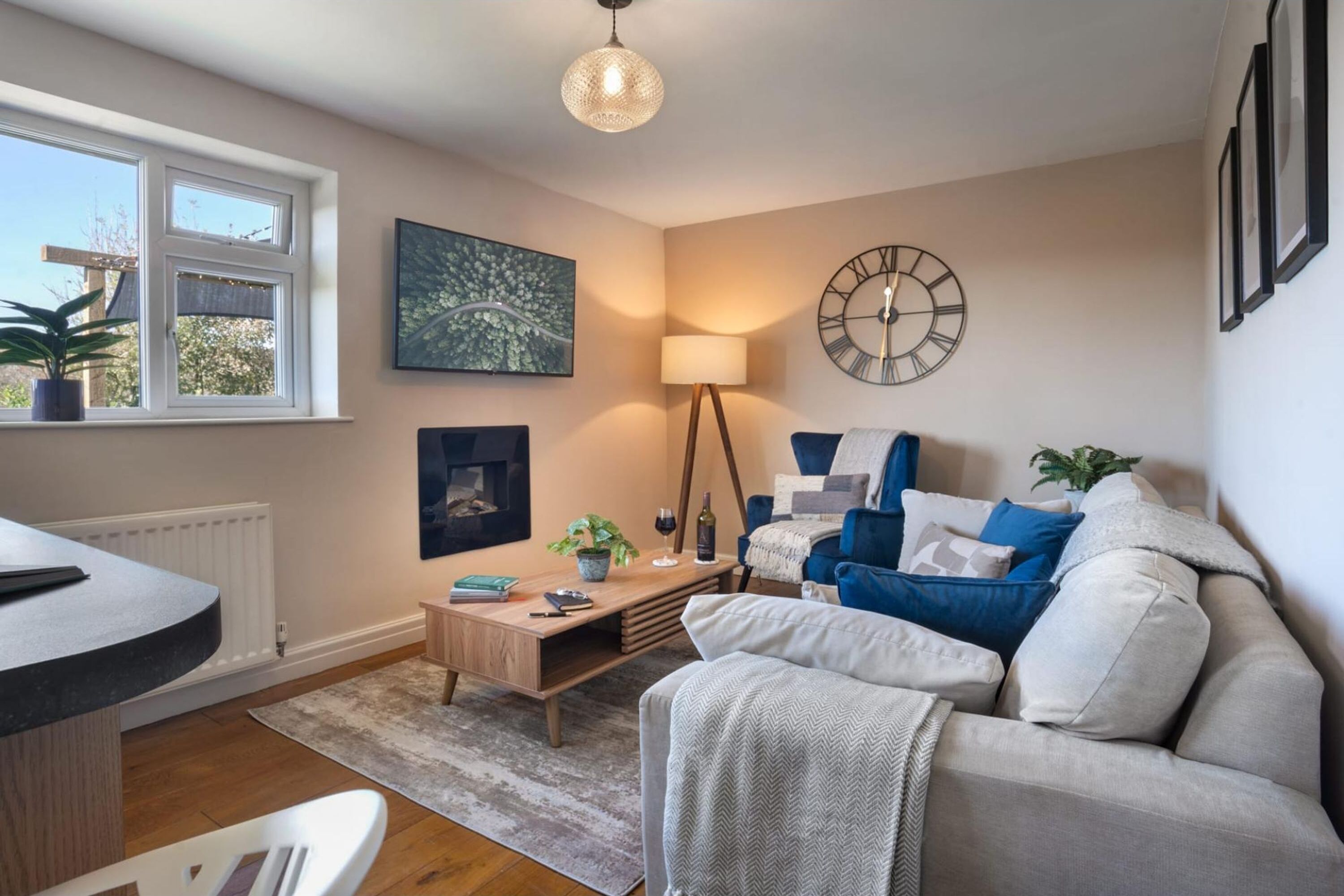 Un salon avec un canapé, une table basse, une lampe, une horloge et une fenêtre ornée de plantes.