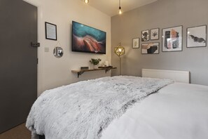A bedroom with a large bed, a fluffy white throw blanket, a mounted TV, a bedside table with a plant, and several framed pictures on the wall. - Windermere Central Hotel - Room 6 (Windermere)