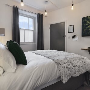 A bedroom with a bed, a window with curtains, a door, a wall-mounted light fixture, and a framed picture. - Windermere Central Hotel - Room 6 (Windermere)