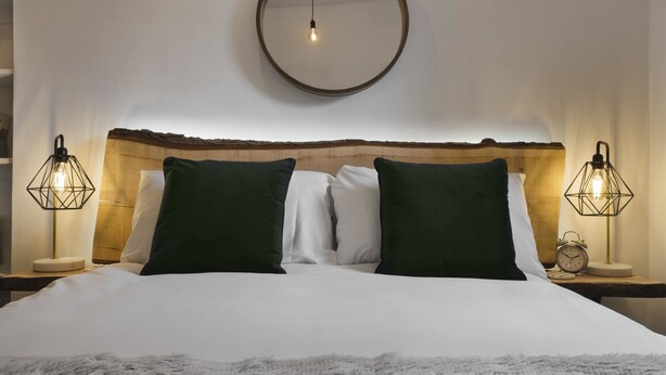 A neatly made bed with a fluffy white blanket, two green pillows, and a wooden headboard. There are two bedside tables with lamps and an alarm clock on one. - Windermere Central Hotel - Room 6 (Windermere)