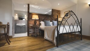 A bedroom with a wooden floor, a bed with a metal frame, a nightstand with a lamp, a brick wall, and a bathroom with a sink and mirror. - Lyth Valley Country House - 9 Rooms (Kendal)