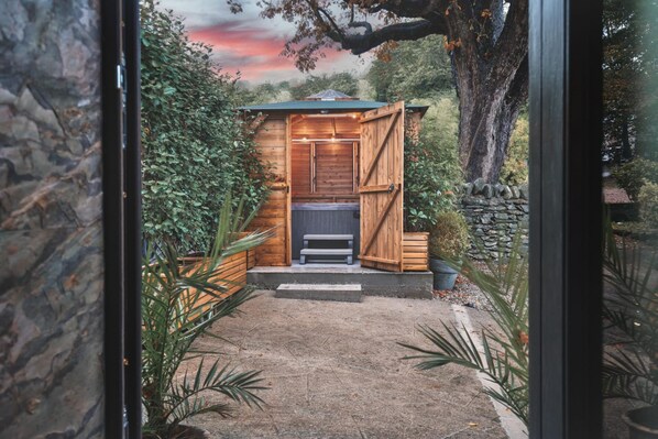 Una cabaña de madera con un banco y una mesa pequeña, rodeada de plantas y un muro de piedra.