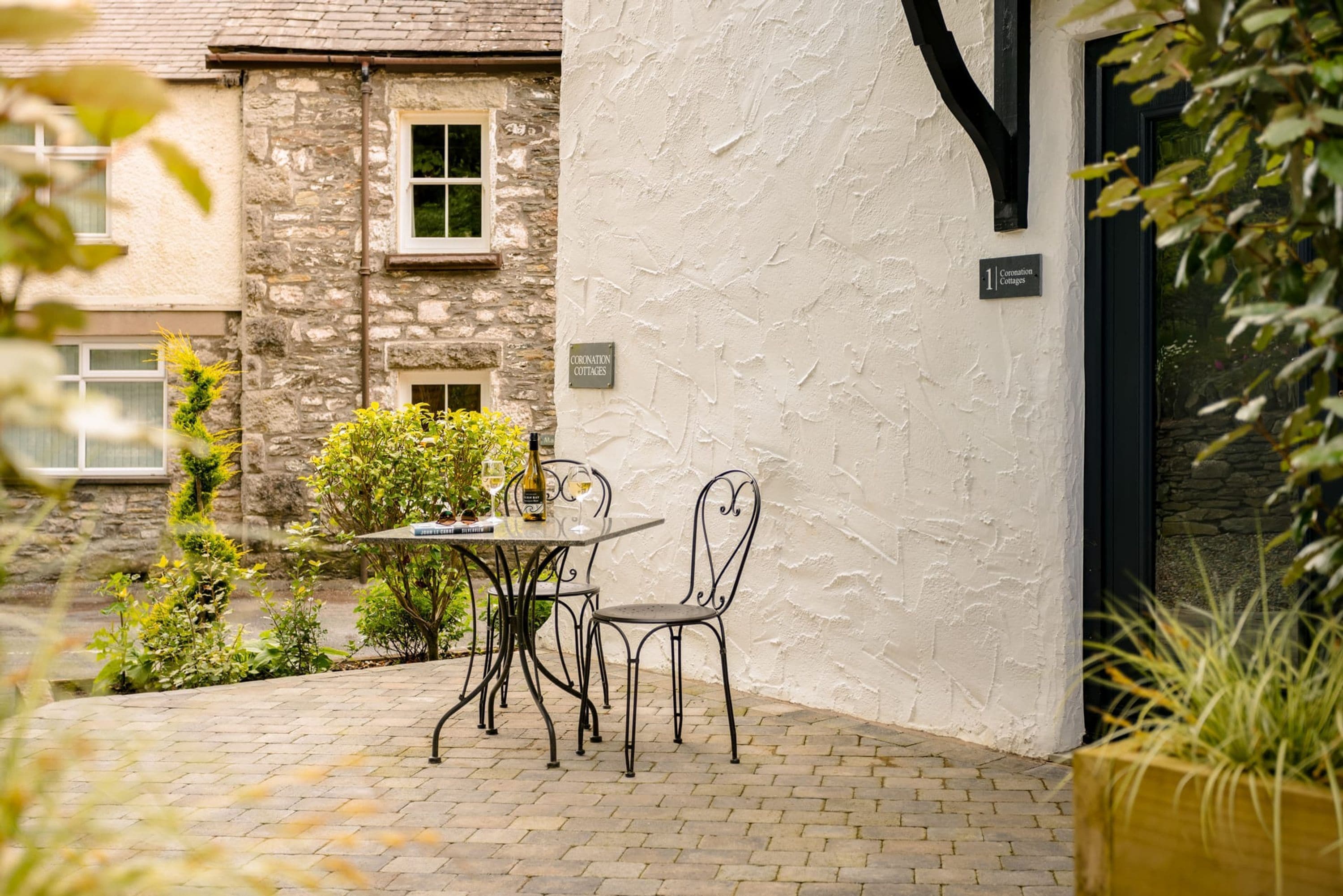 A patio with a table and chairs, a bottle of wine, and a sign indicating '1 Coronation Cottages'.
