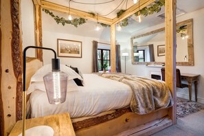 A wooden four-poster bed with a white bedspread and a fur throw. There is a bedside table with a lamp, a mirror, and a framed picture on the wall.