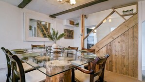 Uma área de refeições com mesa de vidro rodeada por cadeiras de madeira, uma escada de madeira e um espelho na parede.
