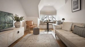 A spacious loft with a high ceiling, a large window, a cozy seating area with a sofa and armchairs, a dining table, and a TV on a cabinet.