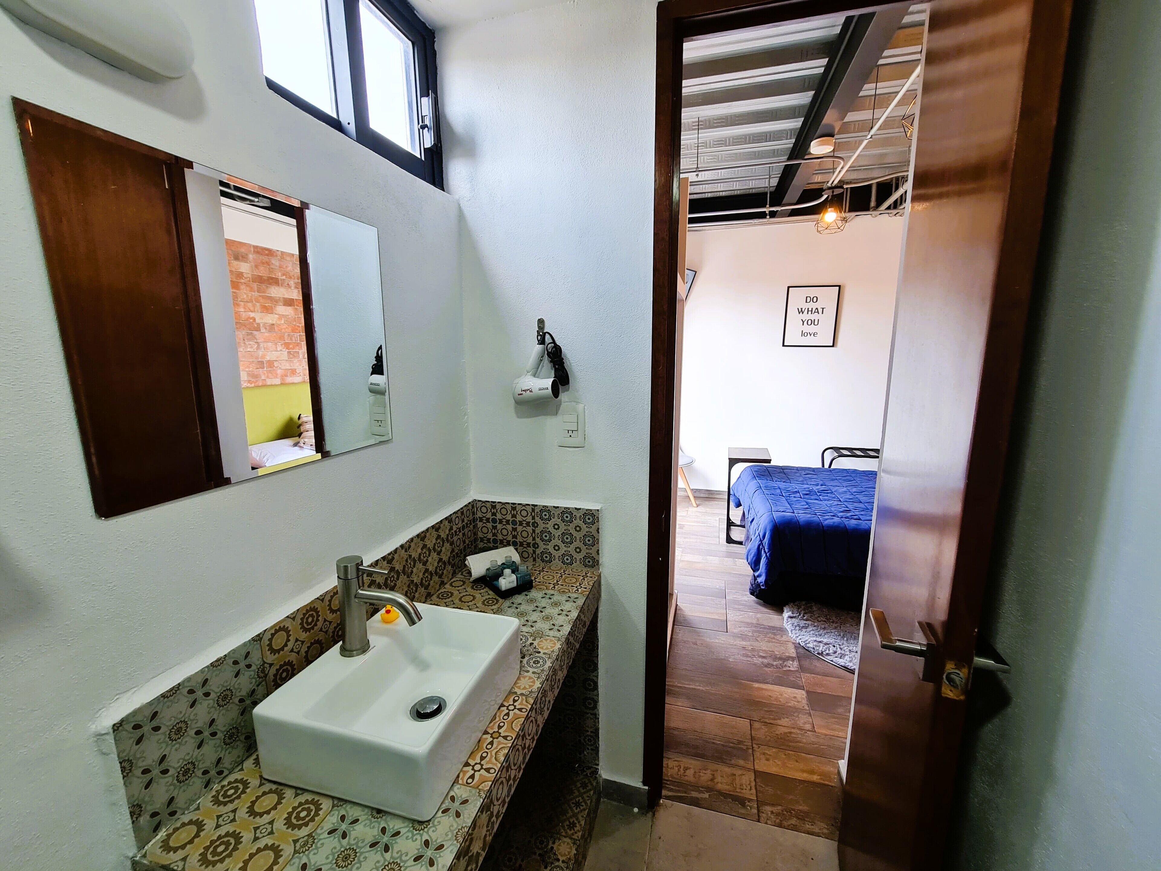Baño con lavamanos, espejo y toallero. Se ve una cama a través de una puerta abierta en la habitación contigua.