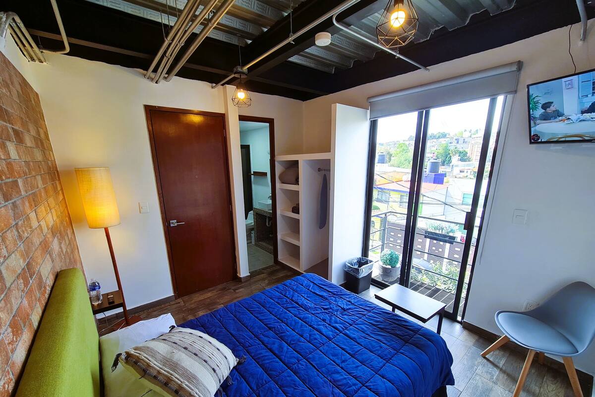Een slaapkamer met een blauwe sprei, een groen kussen, een houten deur, een kleine tafel en een stoel.