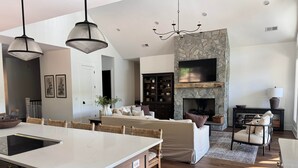 Une cuisine moderne avec un îlot central, un coin repas avec des chaises en osier, un salon avec un canapé et une cheminée, et un mur d’accent en pierre.