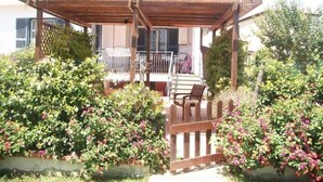 A covered outdoor seating area with a wooden pergola, surrounded by lush greenery and flowering plants.