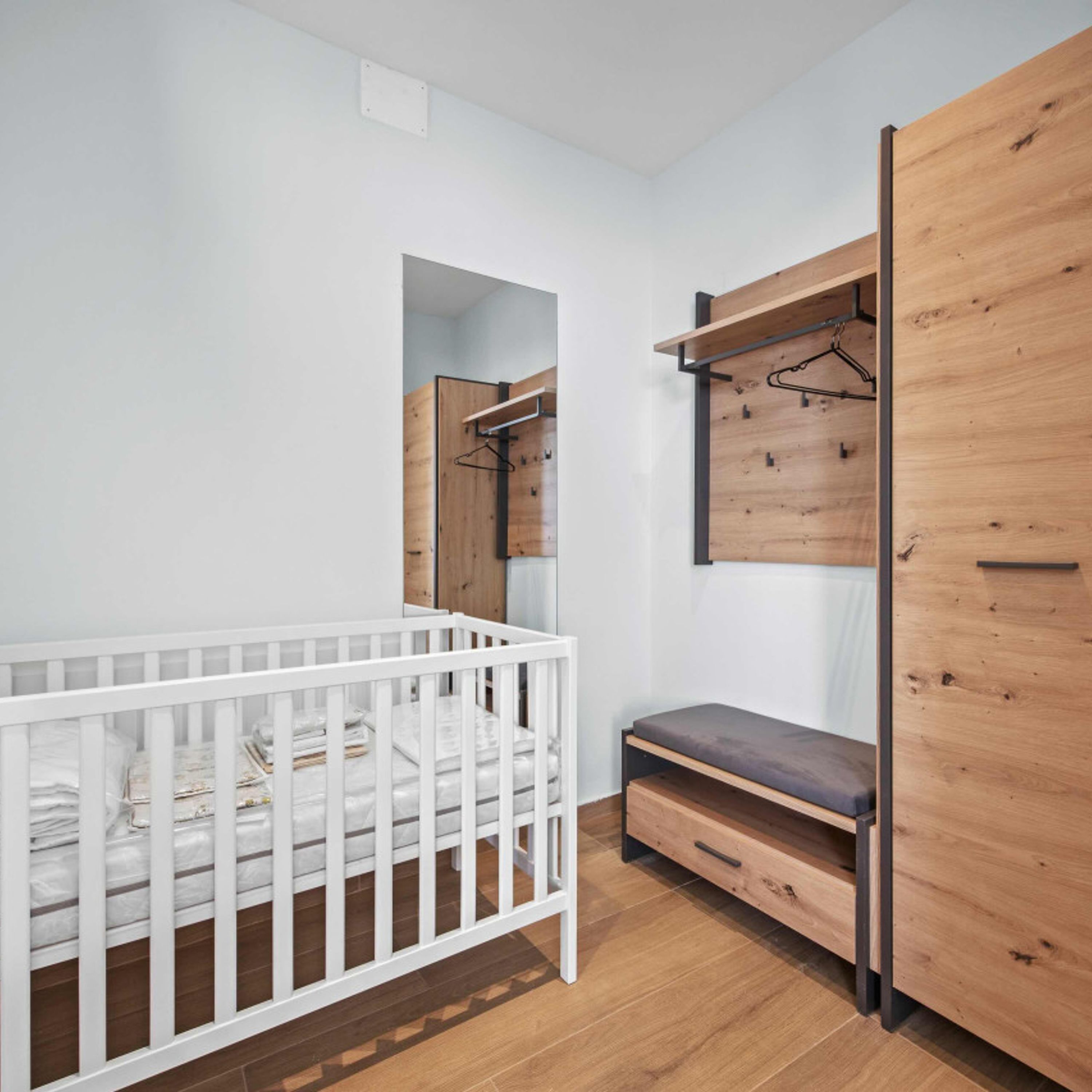 A modern bedroom with a white crib, a wooden wardrobe, and a bench.