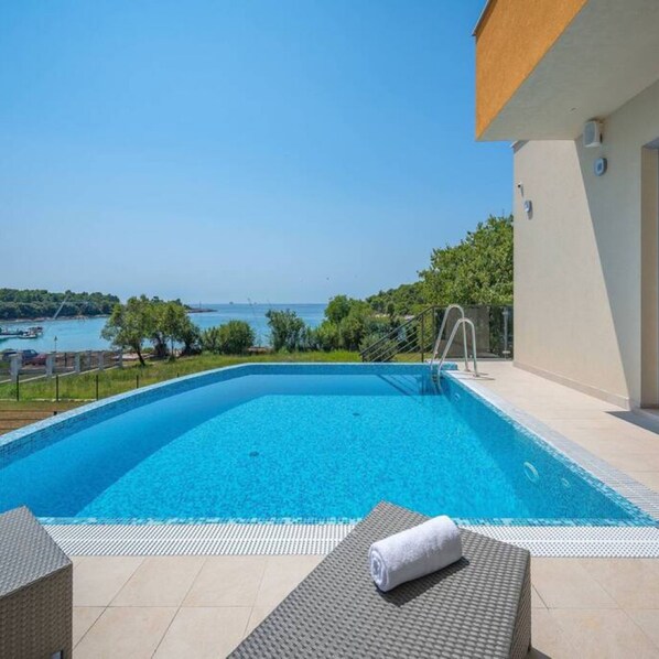 A clear blue swimming pool with a view of the sea and boats, surrounded by a tiled deck and a lounge chair with a towel. - Villa Magnolija in Pula (Pula)