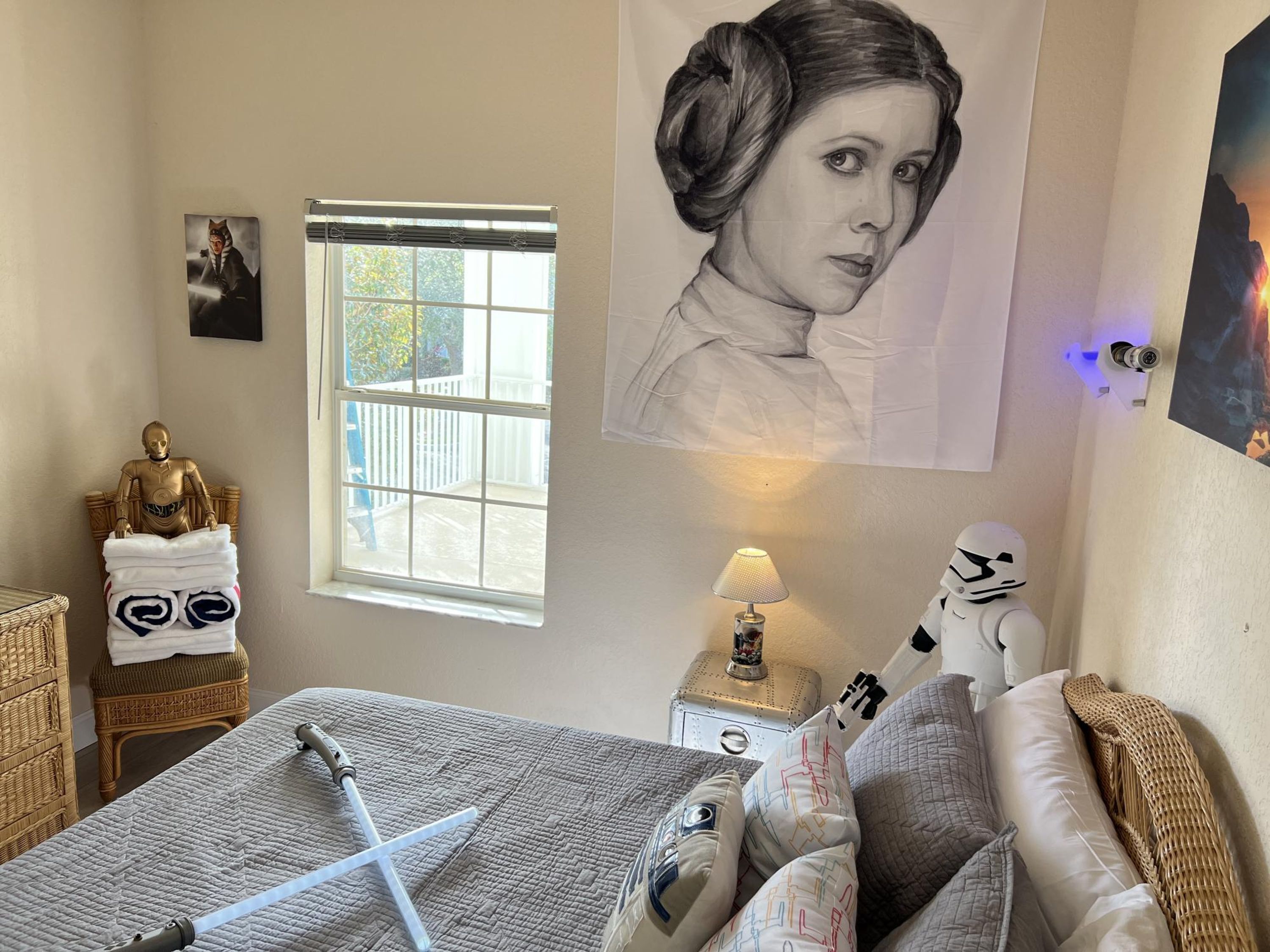 Ett sovrum med en säng, kuddar, en Star Wars-figur, en lampa, ett fönster och en affisch.