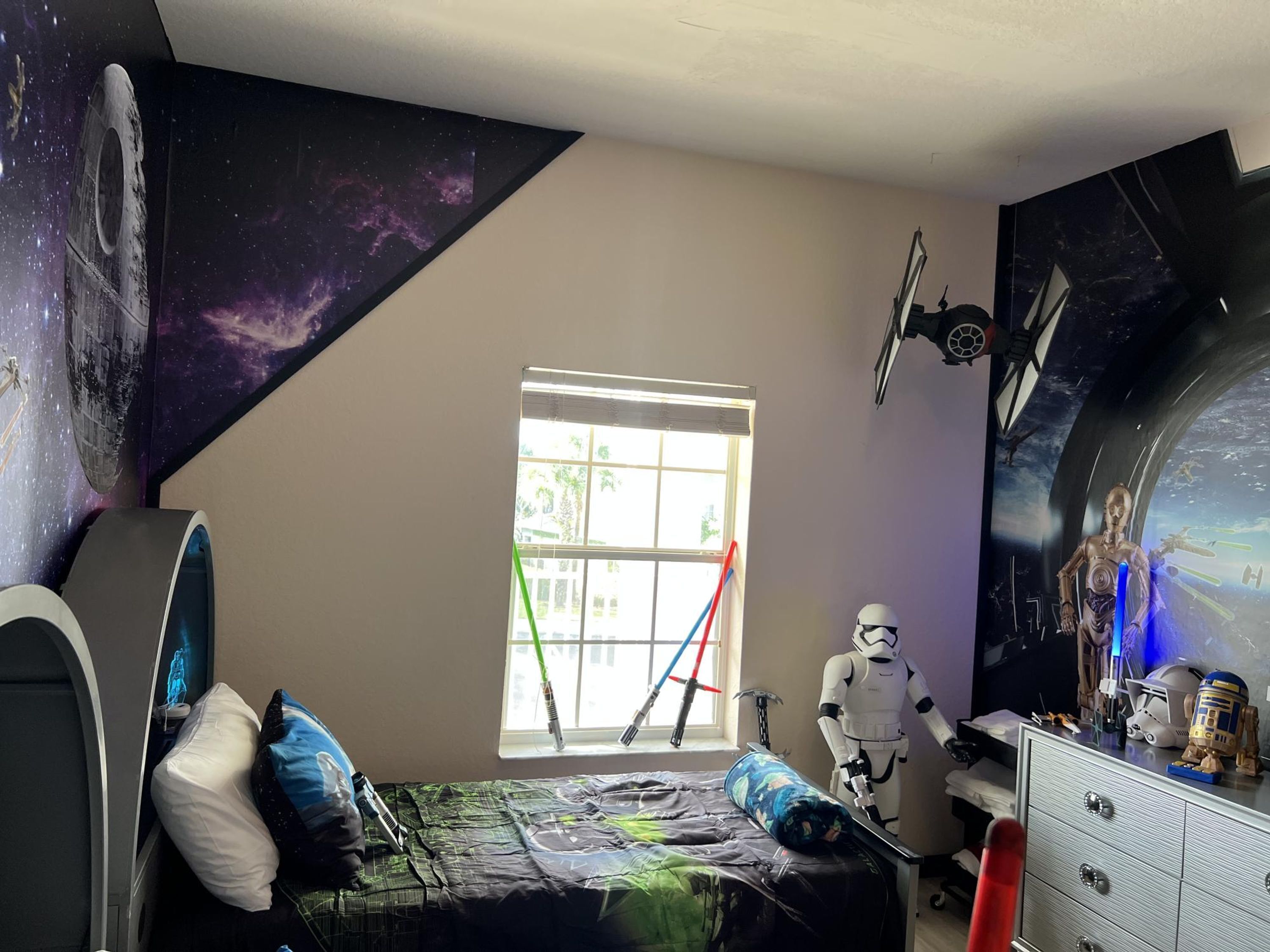 A bedroom with a bed, pillows, a Star Wars-themed wall, a window, and a dresser with toys.