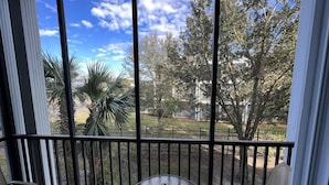 A balcony with a glass table and chairs, overlooking a fenced yard with trees and a clear sky.