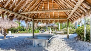 A beachside tennis court with a thatched roof pavilion, surrounded by sand and greenery.