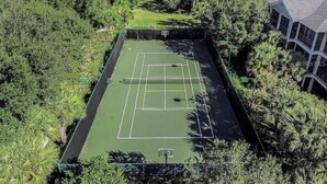 An aerial view of a tennis court surrounded by dense greenery and residential buildings.