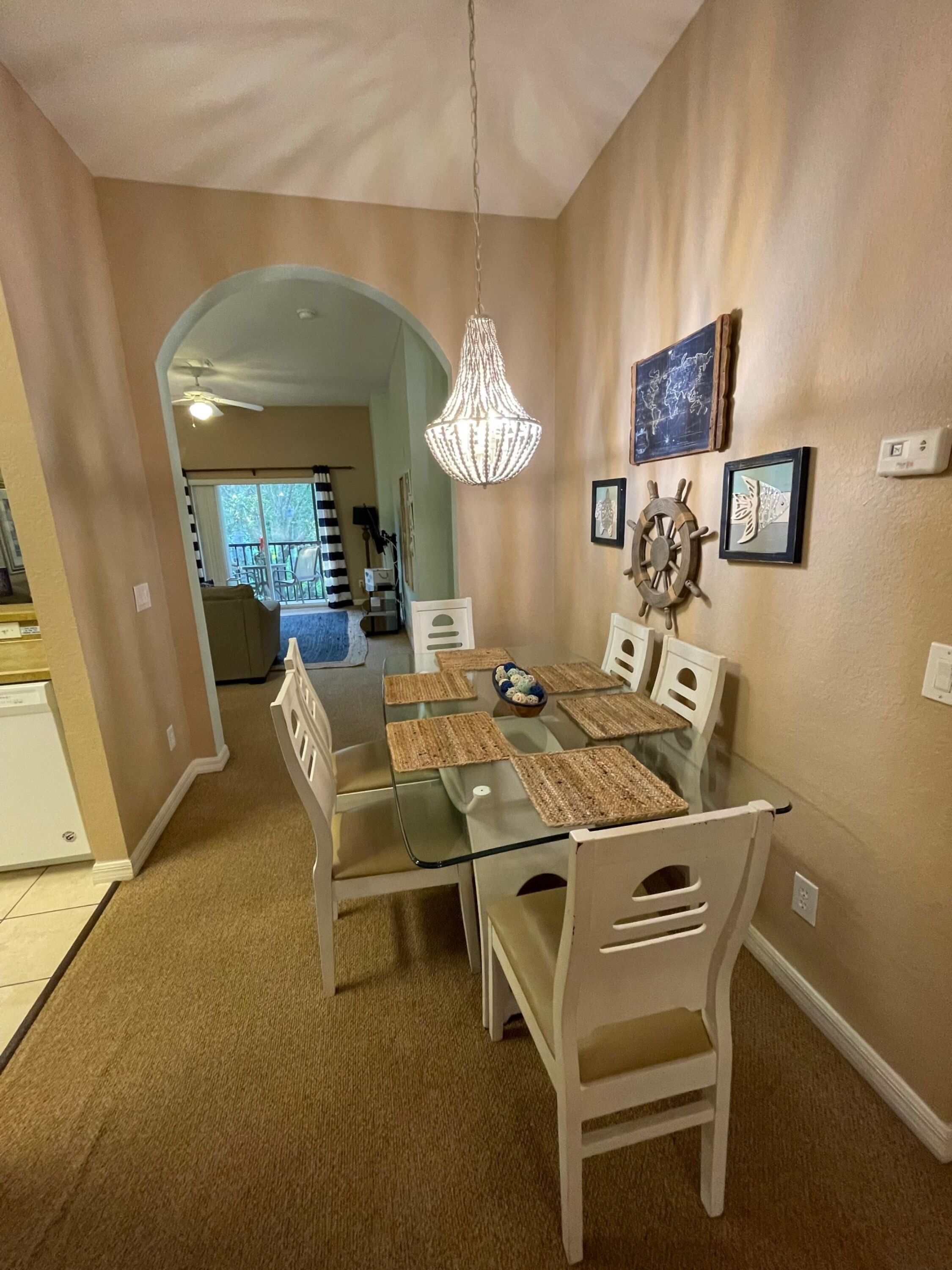 A dining area with a glass table, white chairs, and a chandelier. There are framed pictures on the wall and a view into another room with a couch and a dining table.