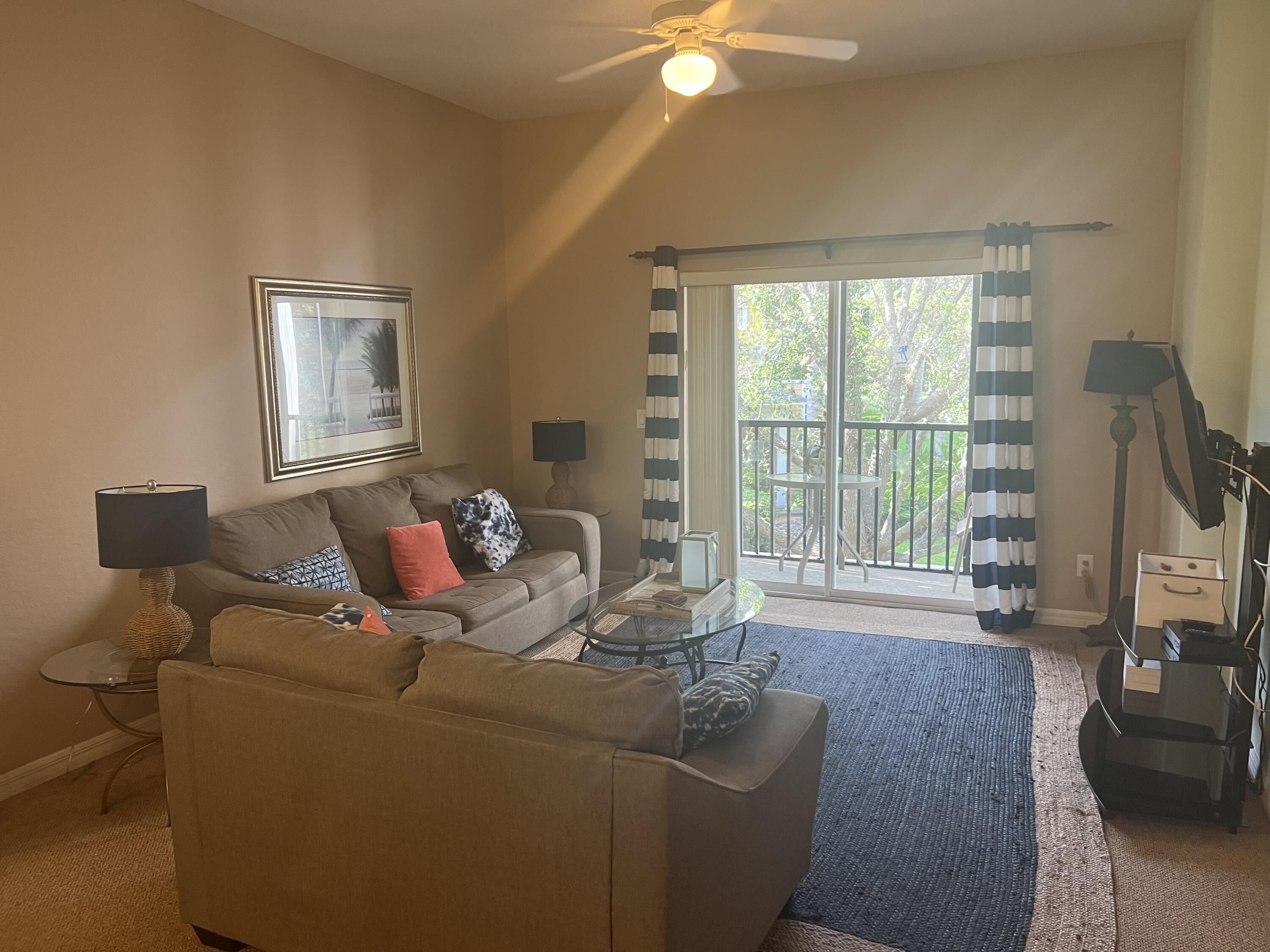 A living room with a sofa, a coffee table, a lamp, and a ceiling fan. There is a window with curtains and a view of greenery outside.