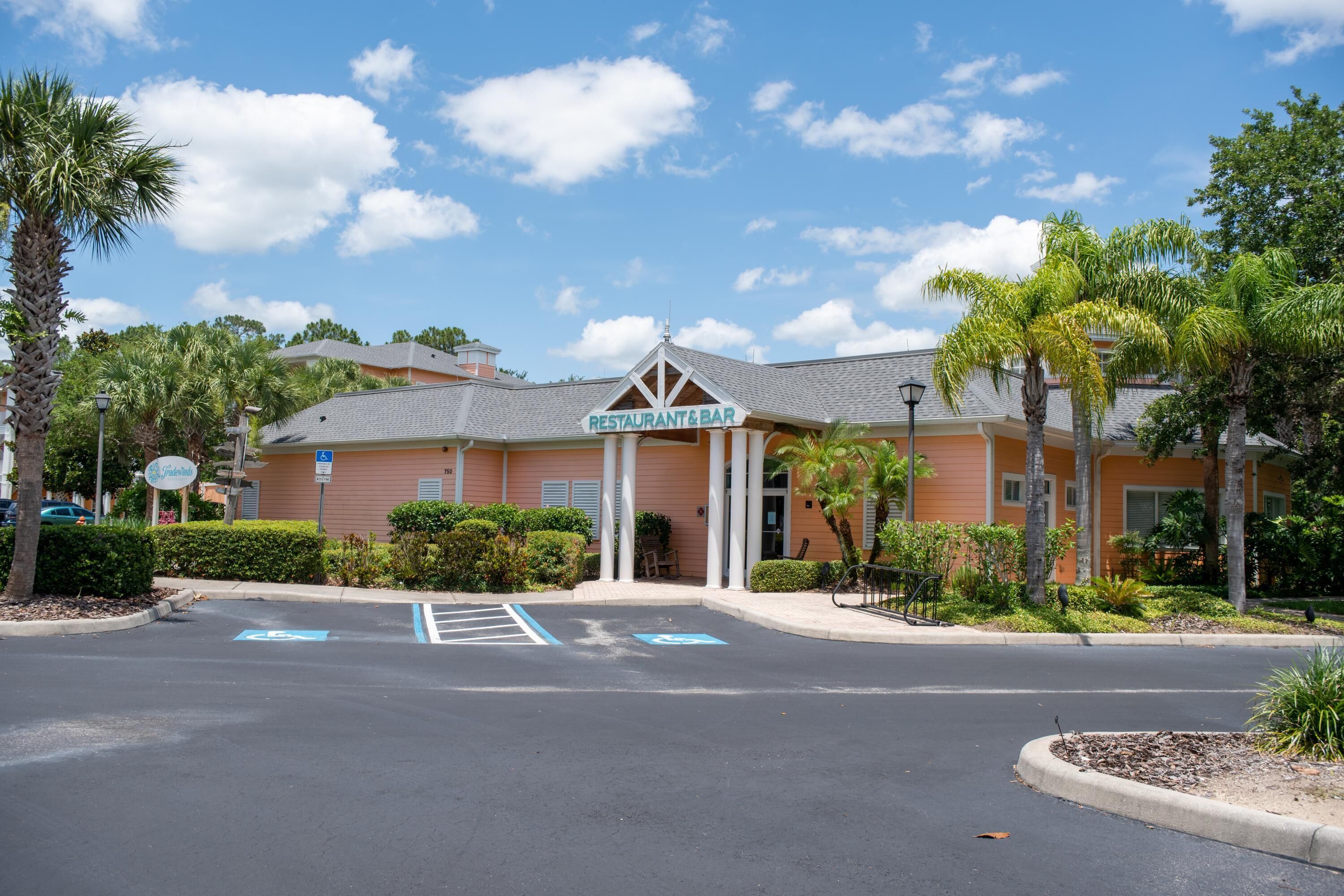 A building with a sign reading "RESTAURANT & BAR" at the entrance, surrounded by palm trees and a parking lot with handicapped parking spaces.