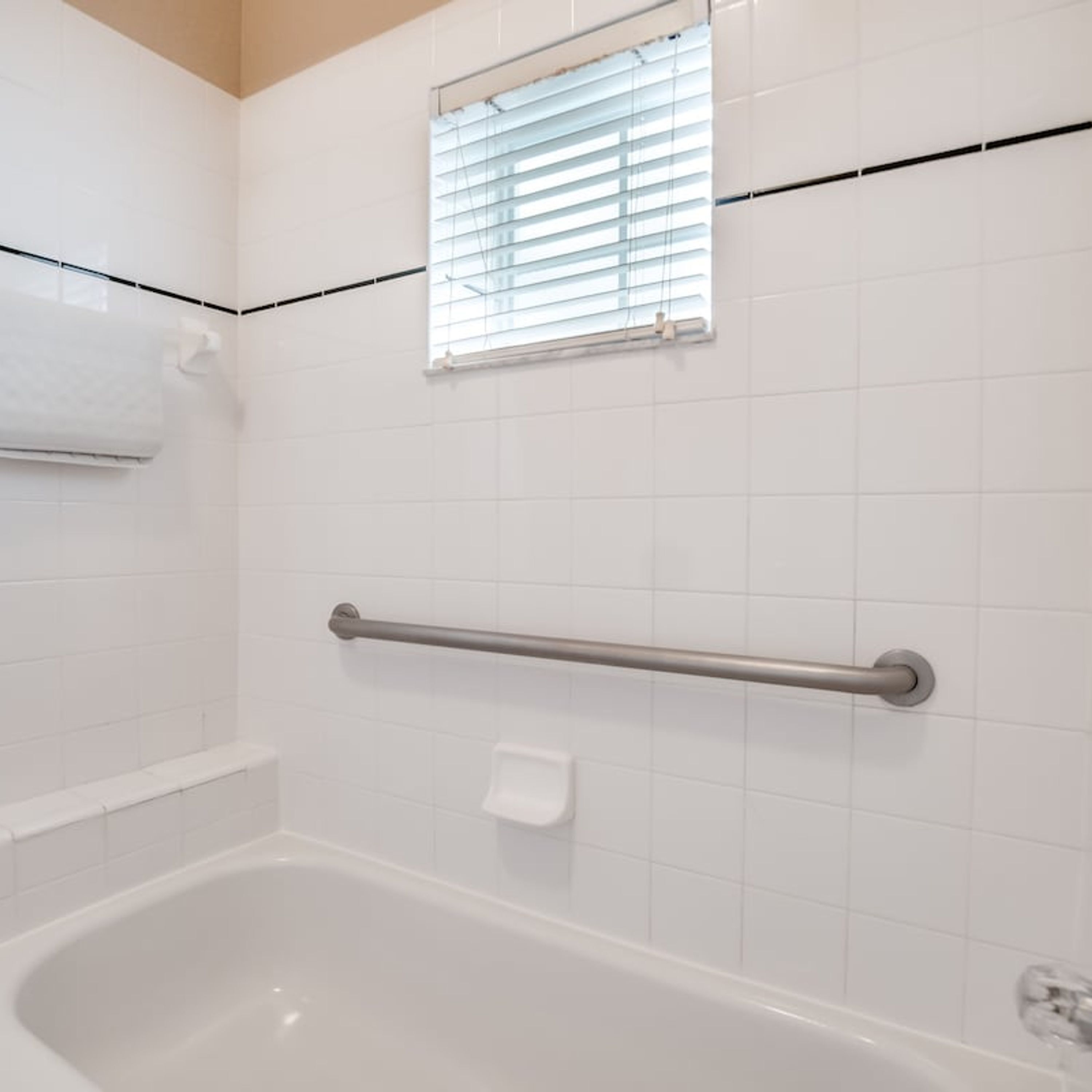 A bathroom with a white bathtub, a window with blinds, and a handrail.