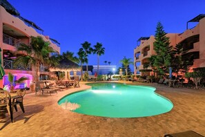 A swimming pool surrounded by lounge chairs and palm trees, with buildings in the background. - El Pueblo Condominiums by Rocky Host  (Puerto Peñasco)