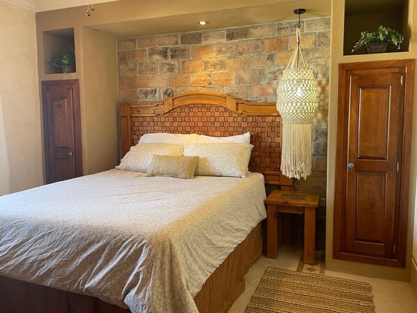 A bedroom with a brick-patterned headboard, a wooden nightstand, a hanging macrame light fixture, and a wooden wardrobe. - El Pueblo Condominiums by Rocky Host (Puerto Peñasco)