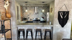 Cocina compacta con una isla central, tres taburetes y un adorno de macramé colgado.