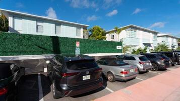 Un parking où plusieurs voitures sont garées en rangées, avec une haie verte et des bâtiments en arrière-plan.