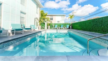 A swimming pool with a tiled edge, surrounded by lounge chairs and a green hedge.